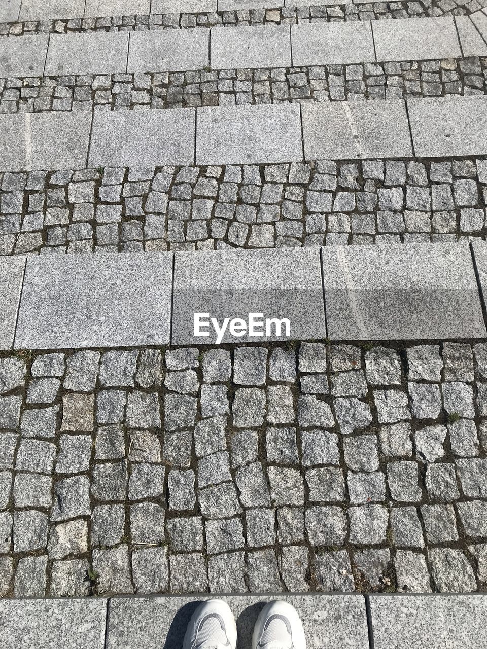 wall, road surface, cobblestone, flooring, low section, pattern, day, human leg, high angle view, brick, footpath, shoe, street, outdoors, floor, one person, textured, art, directly above, stone, footwear, standing, city, paving stone, full frame, nature, architecture, iron, close-up, stone wall, tile, sunlight, asphalt, soil