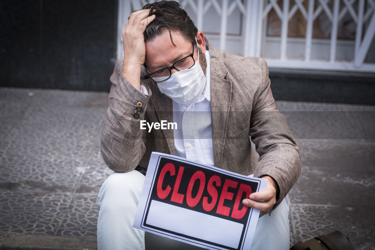 Sad man with head in hands wearing mask sitting on footpath