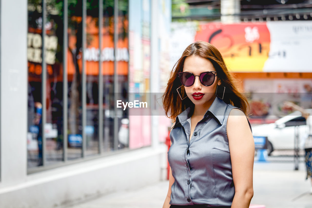 Portrait of beautiful young woman wearing sunglasses on footpath in city