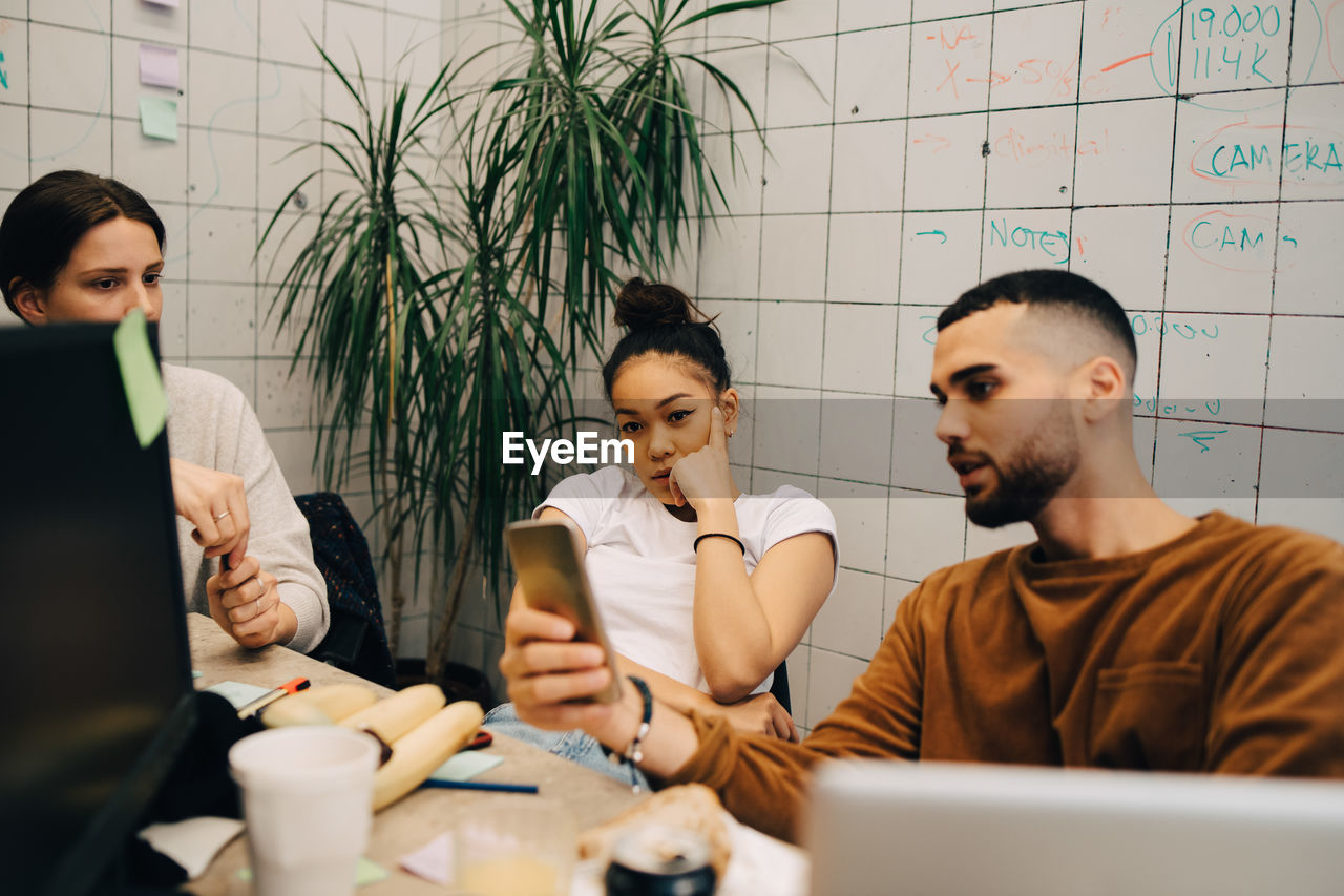 Young male hacker sharing smart phone with female colleagues while sitting at creative office