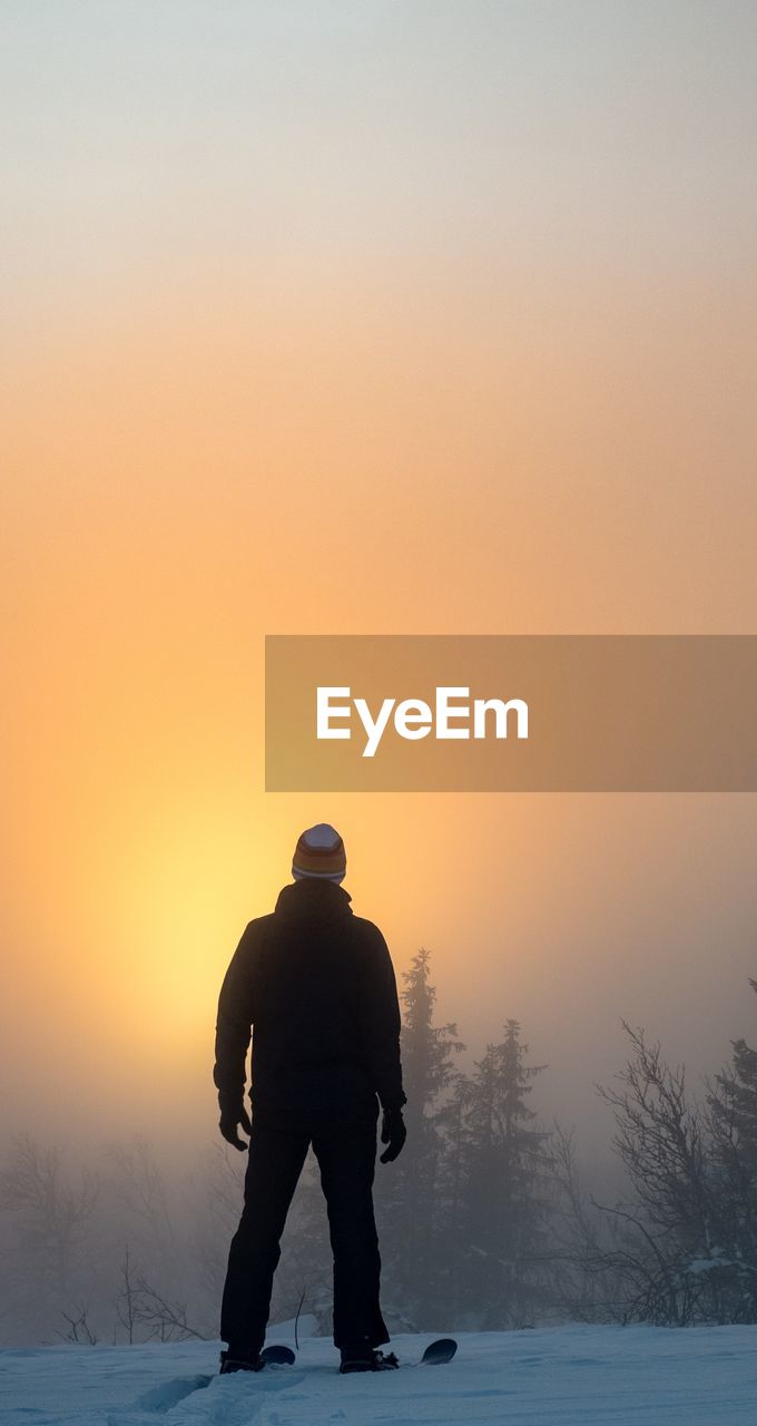 Rear view of silhouette mid adult man standing on snow covered field against clear sky during sunset