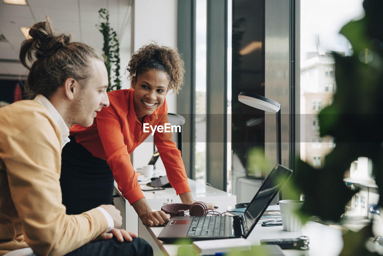 Smiling businesswoman leaning on desk while looking at businessman sitting at office