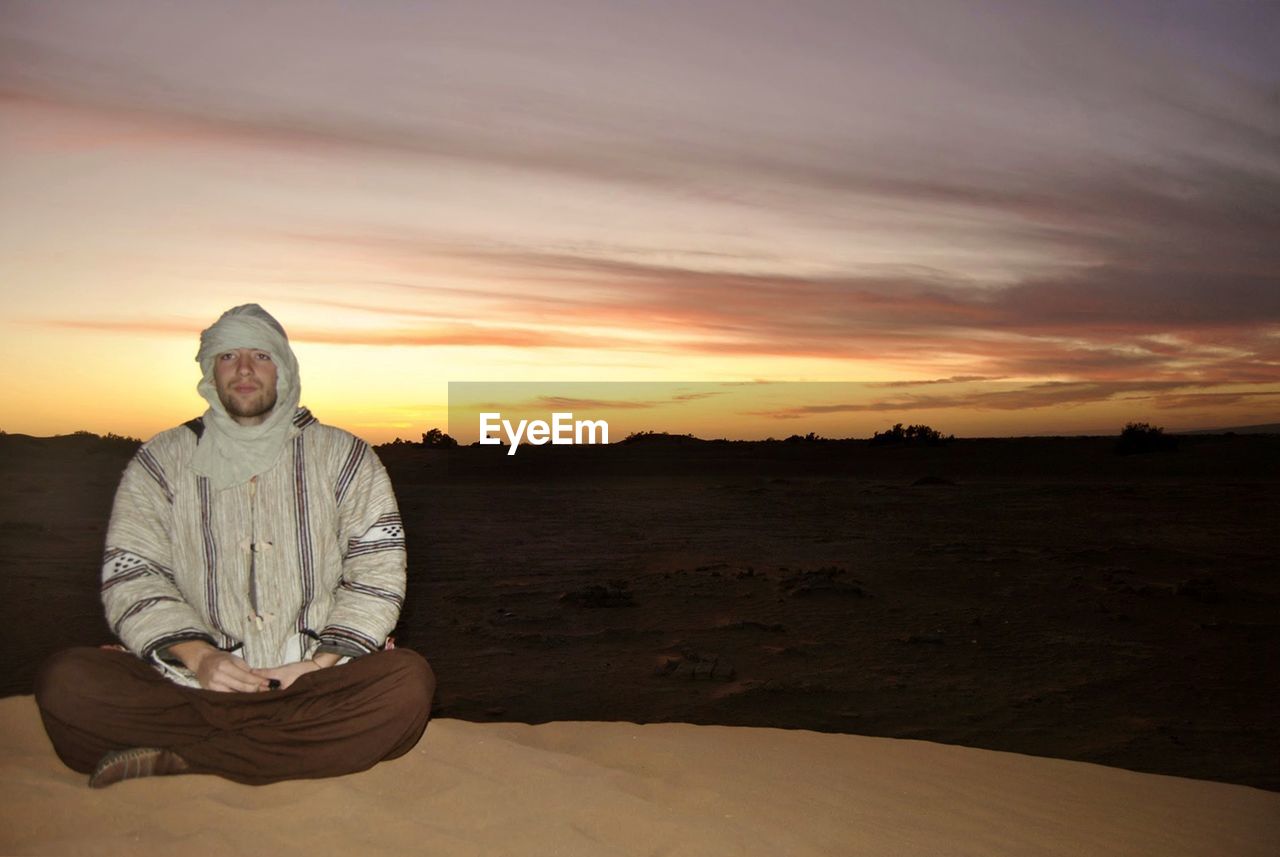 Portrait of man sitting on sand in desert against sky during sunset