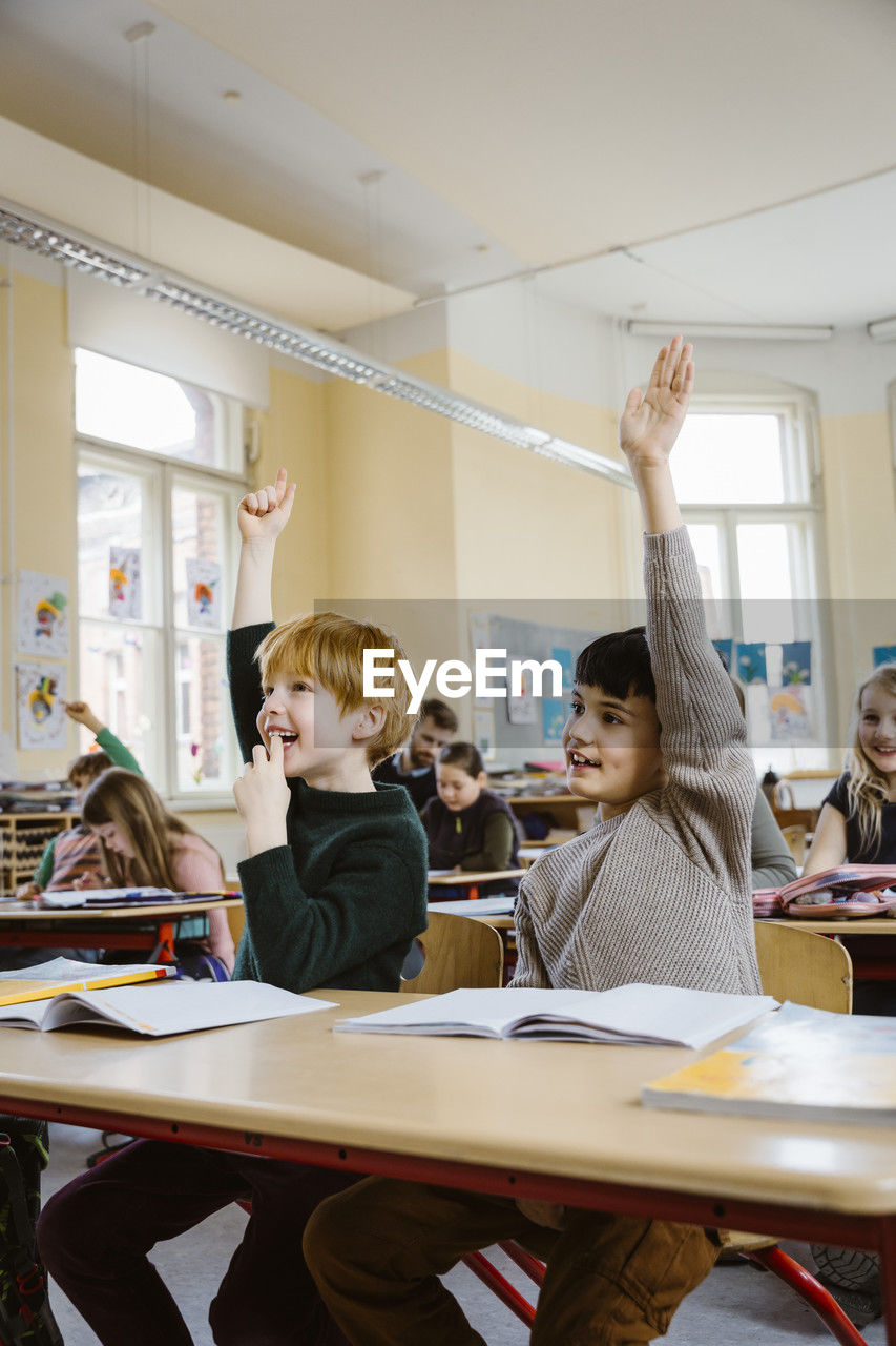 Smiling male pupils raising hands while answering in classroom