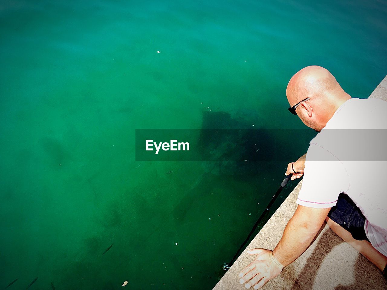 High angle view of mature man fishing in sea while kneeling on retaining wall