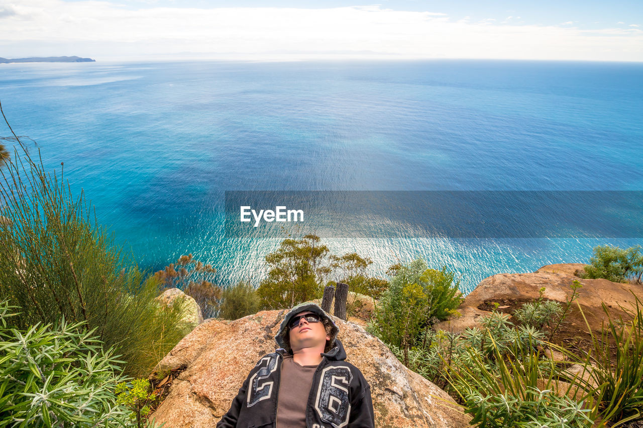 High angle view of young man lying on rock by sea against sky