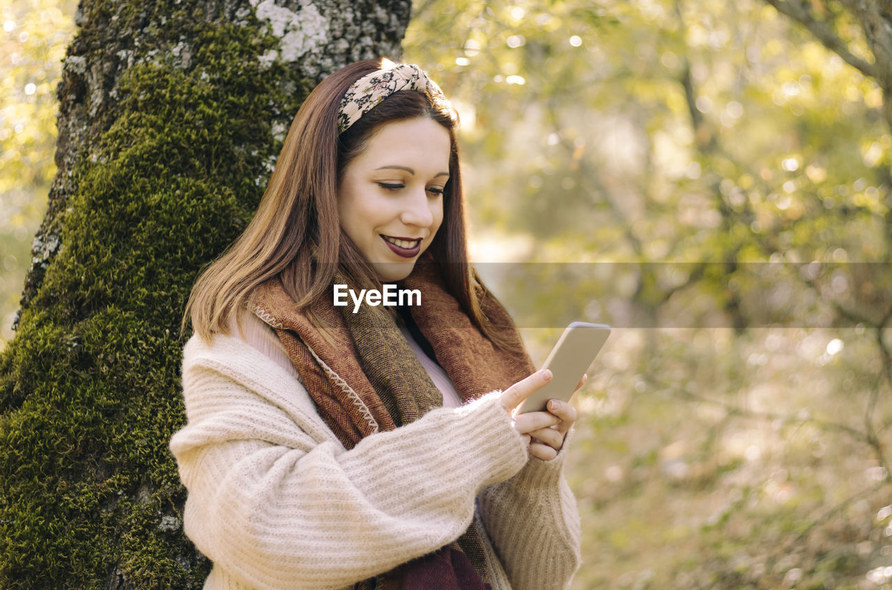 Smiling young woman, leaning against a tree, using her mobile phone
