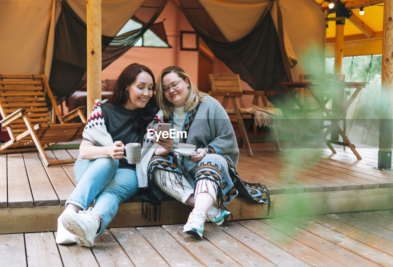 Two young women friends using mobile phone relaxing in glamping in the woods