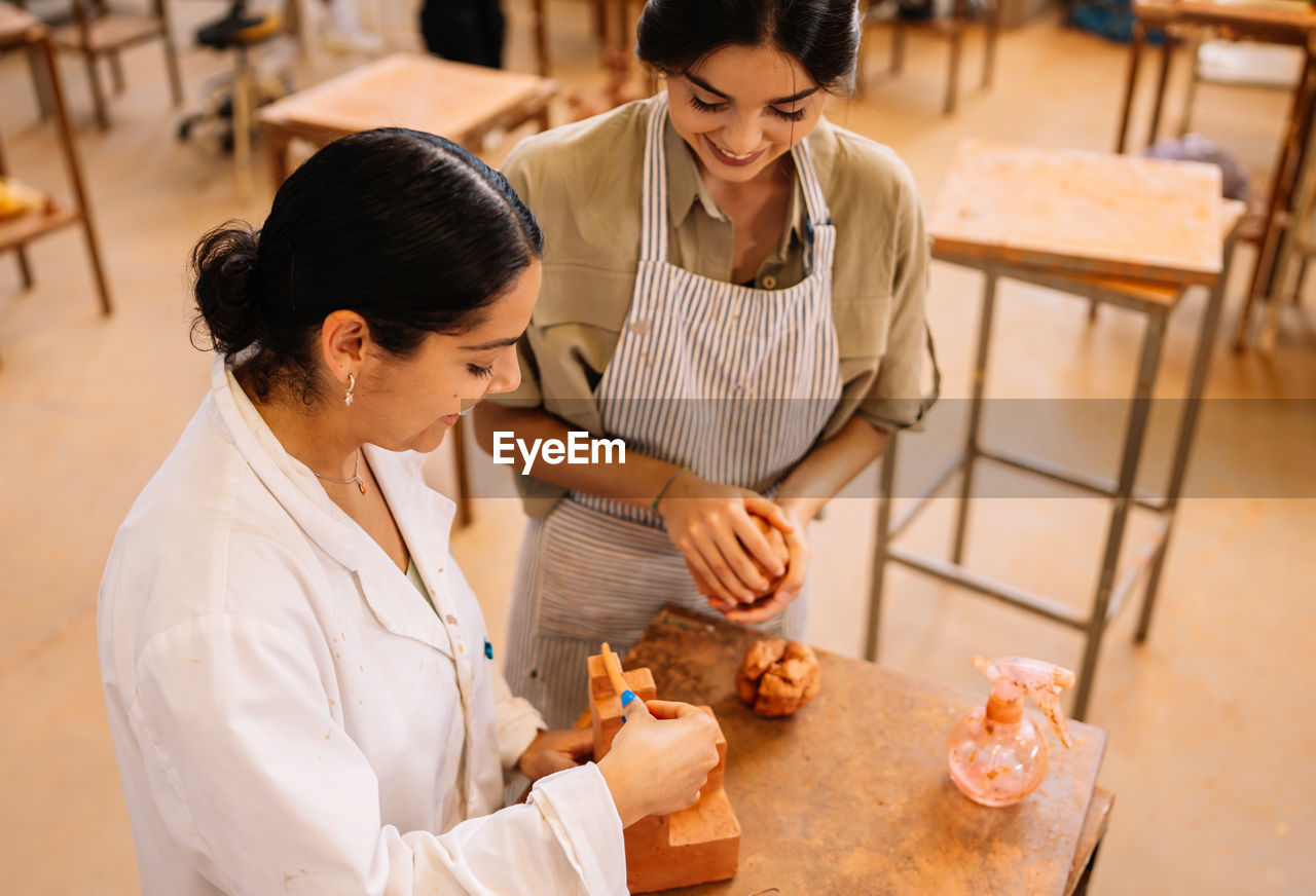 From above merry hispanic girlfriends smiling and creating geometric shape from clay on table in spacious workshop