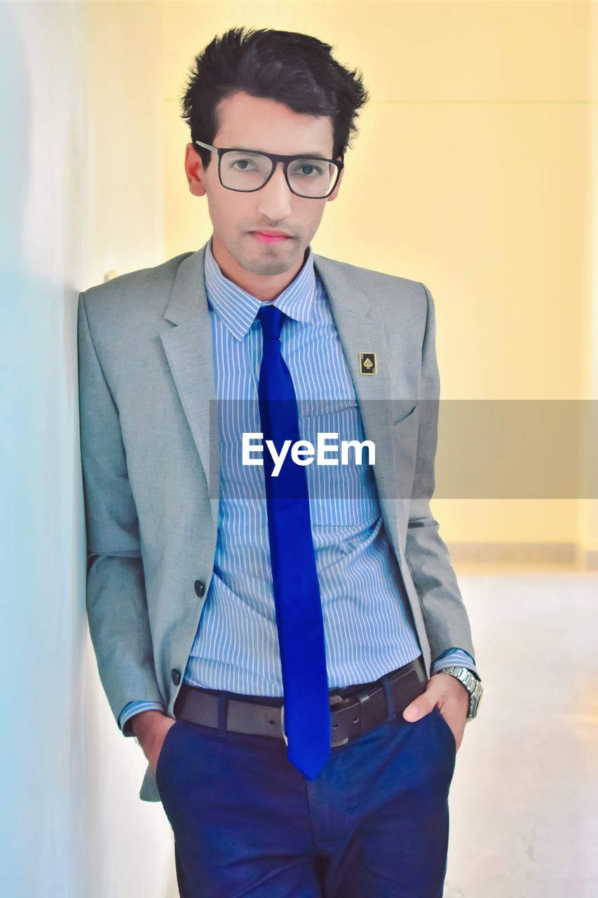 one person, businessman, eyeglasses, business, men, glasses, adult, blue, indoors, spring, formal wear, portrait, young adult, standing, blazer, necktie, front view, clothing, corporate business, looking at camera, menswear, hands in pockets, tie, three quarter length, office, button down shirt, dress shirt, businesswear, fashion, serious, looking, shirt, occupation, professional occupation, business finance and industry, white-collar worker, cool attitude, shirt and tie, black hair, brown hair, trousers, outerwear