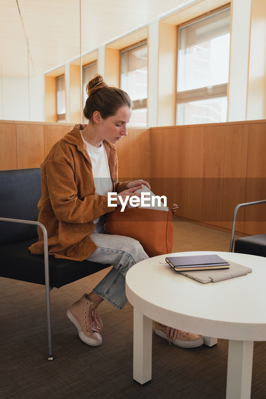 Full body young female student in casual clothes with hair bun taking stationery from bag while sitting on chair near table and preparing for studies in university library