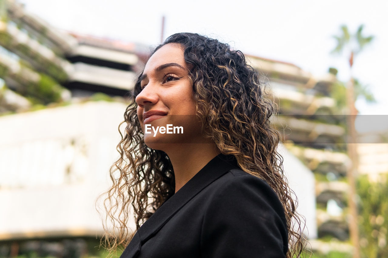 From below side view self assured young hispanic businesswoman in stylish black jacket with curly hair looking away on blurred background of city street