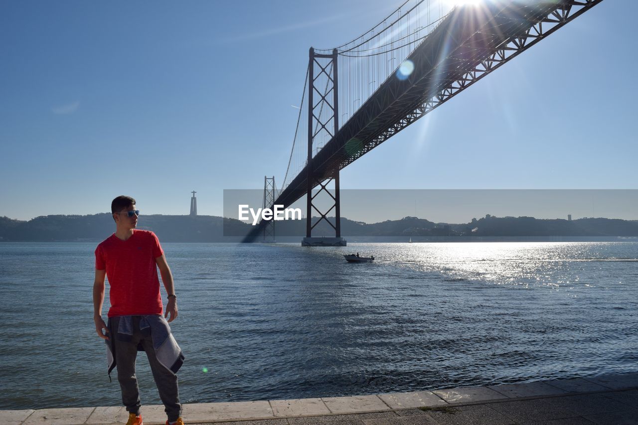 Young man standing on riverbank against clear sky during sunny day