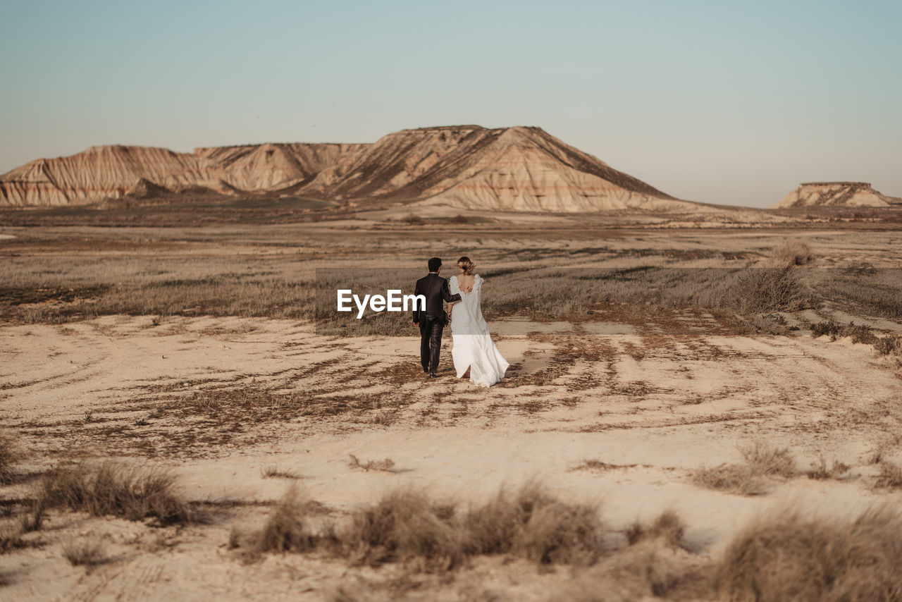 Back view of unrecognizable bride and groom in elegant clothes walking on arid plain towards mountains during wedding celebration in bardenas reales natural park in navarra, spain