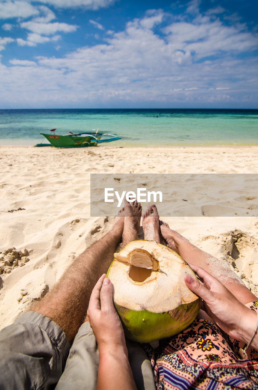 Low section of couple with coconut water relaxing at beach on sunny day