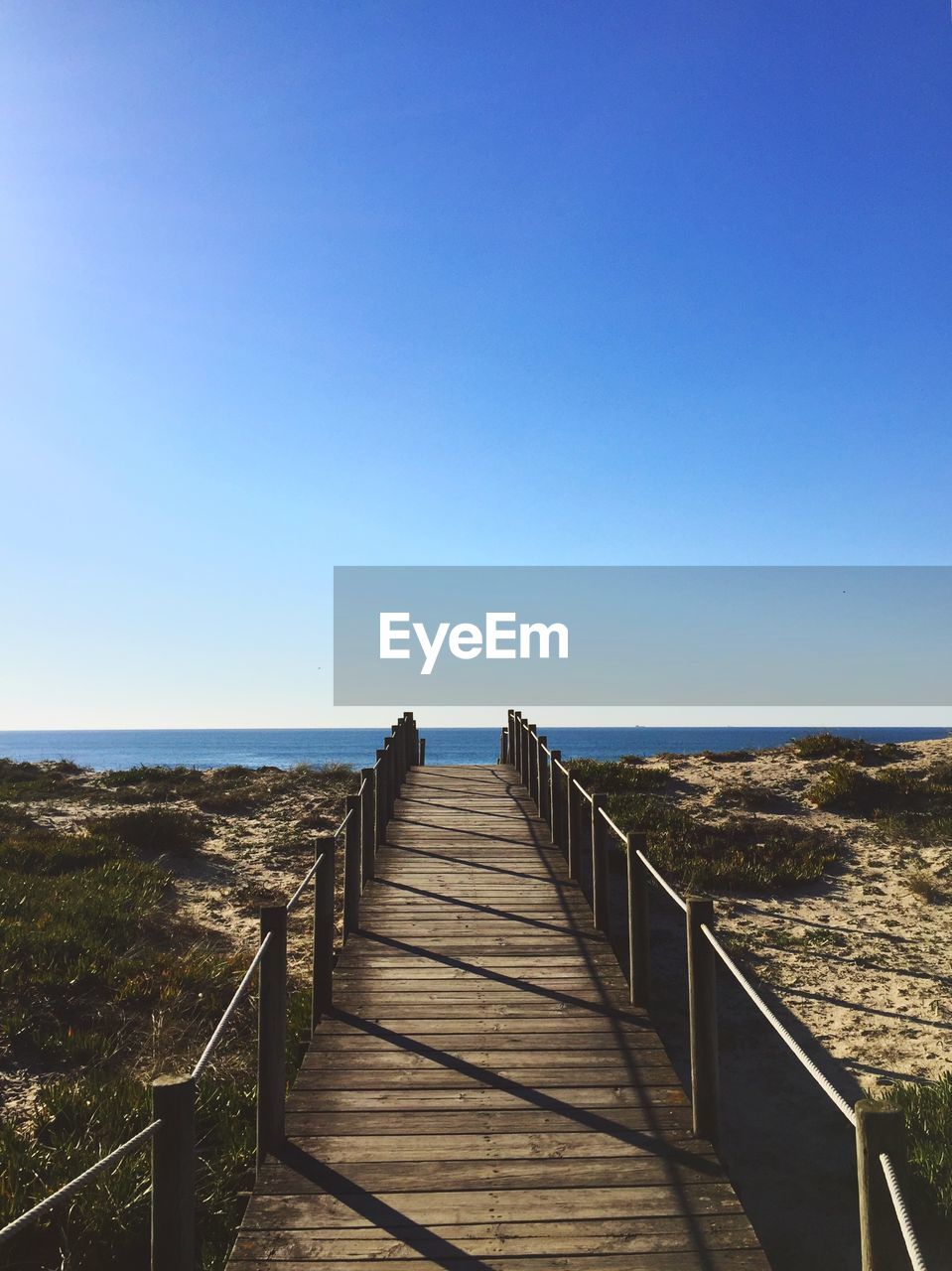 Wooden walkway leading towards sea against clear blue sky