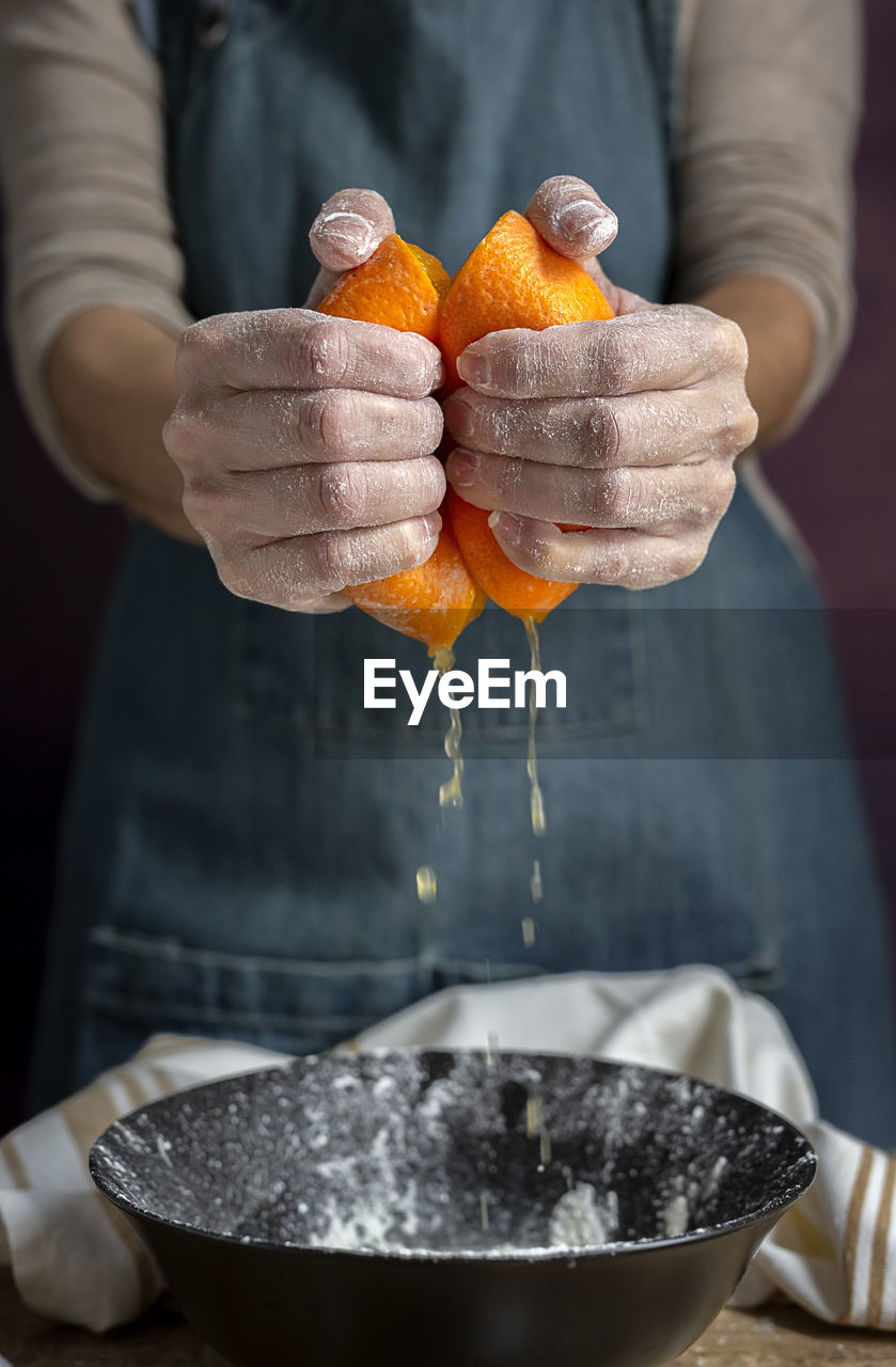 Crop hand of unrecognizable woman in apron squeezing fresh juicy cut orange over bowl while preparing dough at table