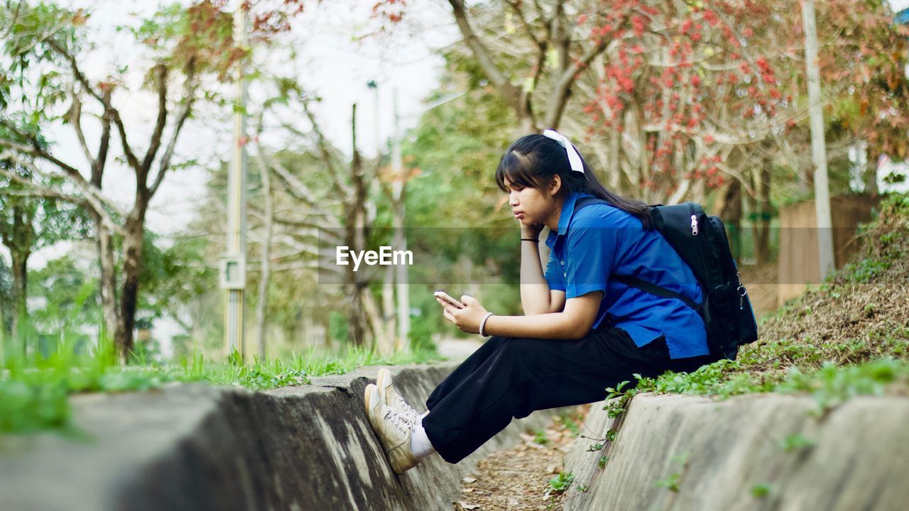 Young woman with backpack using mobile phone while sitting against plants in park