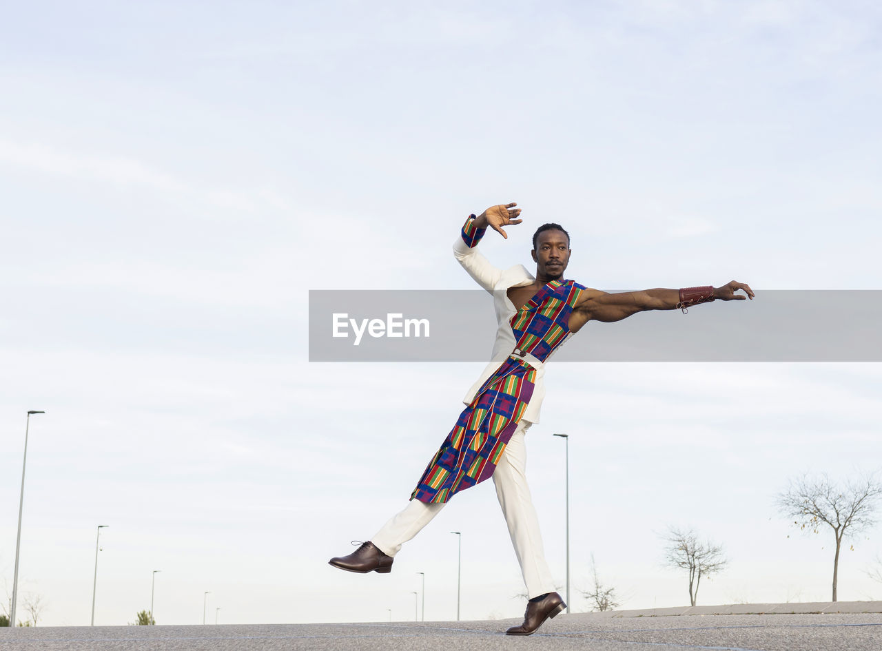 Side view of african american male dancer in trendy outfit performing energetic movements against blue sky