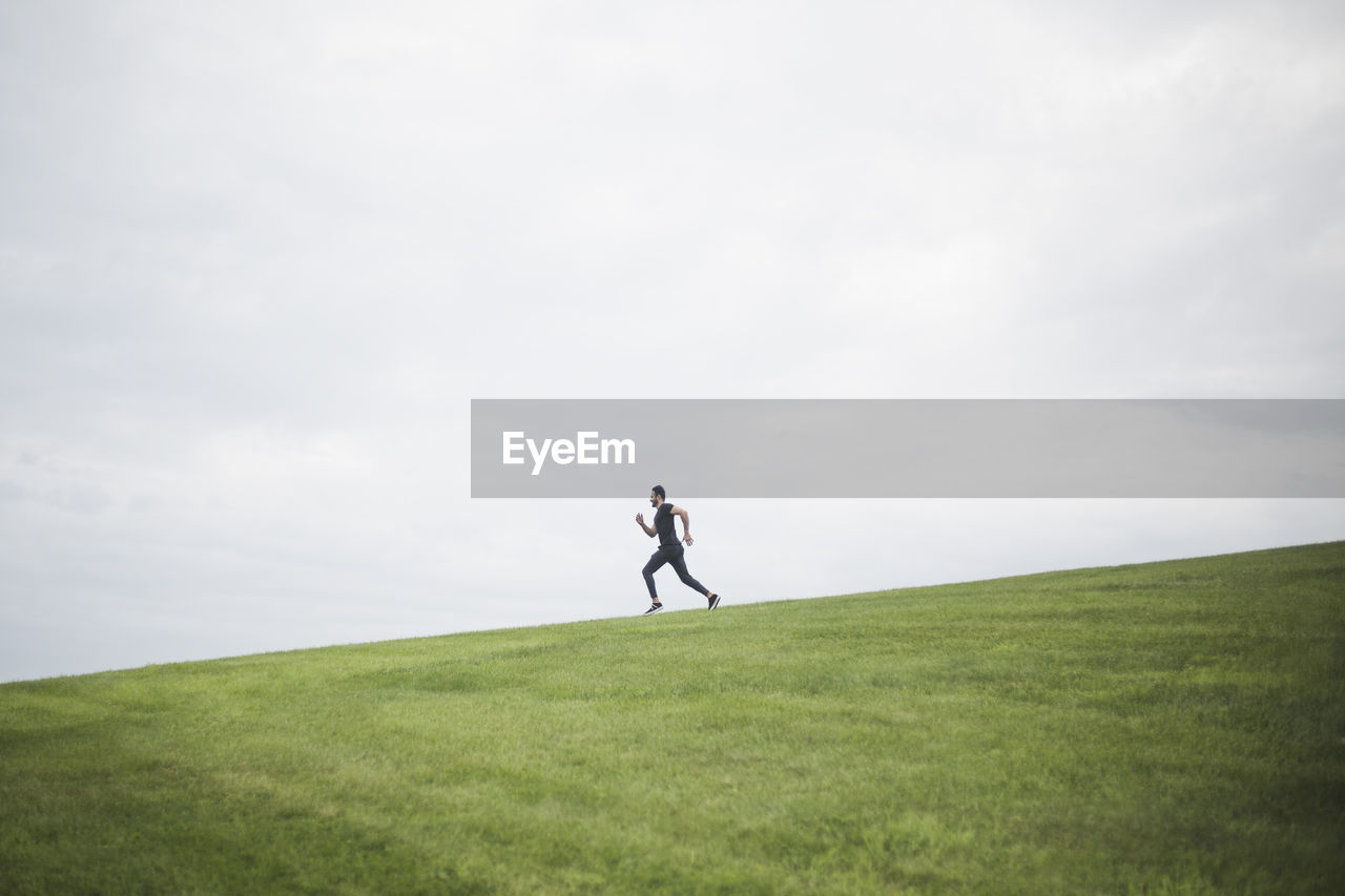 Side view of young man running on field against cloudy sky