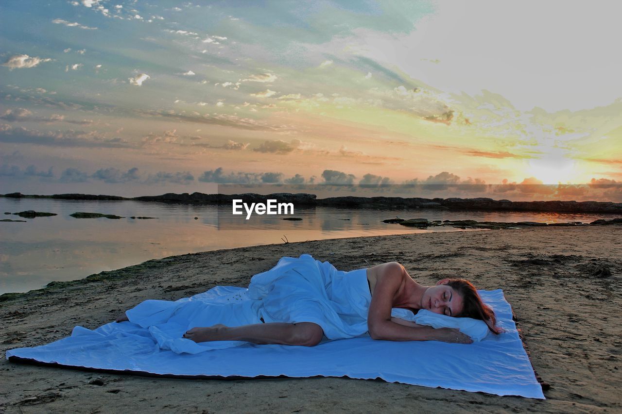 Woman lying on bed at beach against sky during sunset