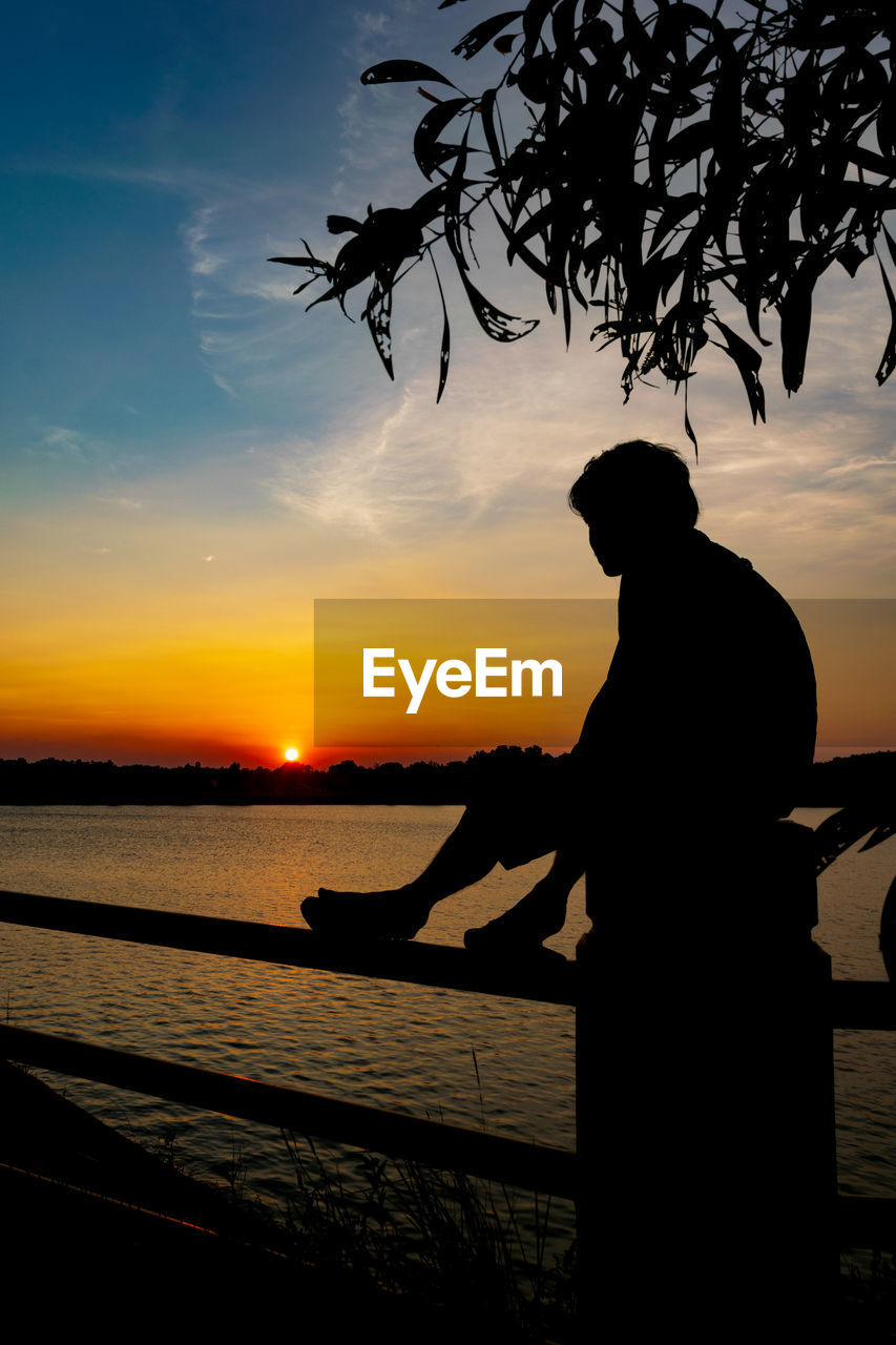 Silhouette man sitting on railing by sea against sky during sunset