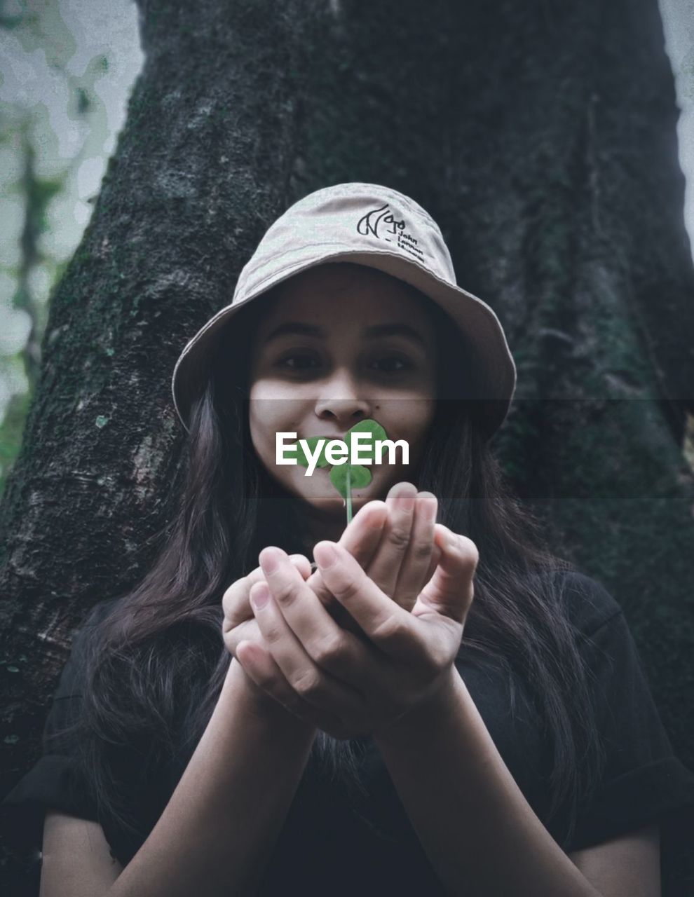 one person, portrait, hat, tree, women, looking at camera, plant, child, front view, nature, holding, childhood, black, clothing, fashion accessory, tree trunk, adult, young adult, emotion, outdoors, trunk, cap, person, female, forest, day, long hair, cute, lifestyles, casual clothing, hand, smiling, leisure activity, green, human face, portrait photography, happiness, headshot, hairstyle