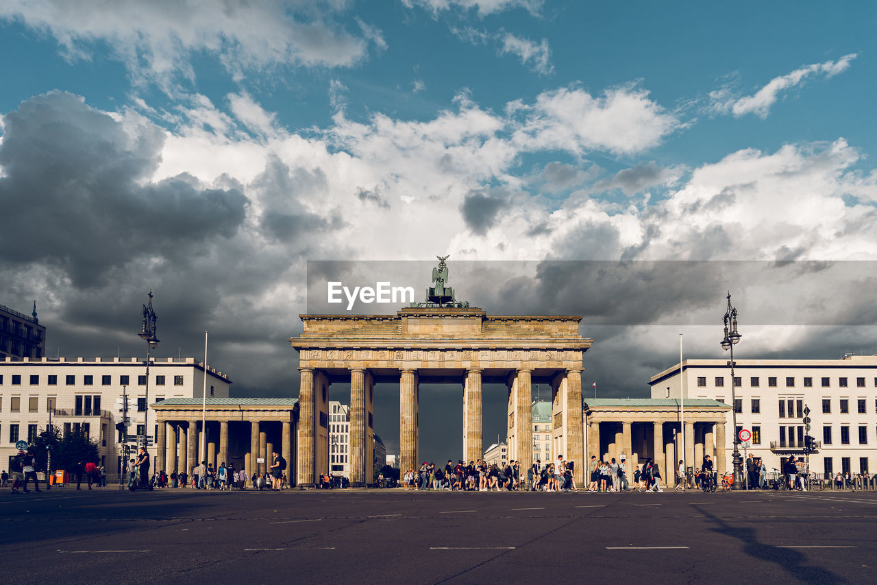 Group of people in front of brandenburg gate and buildings against cloudy sky