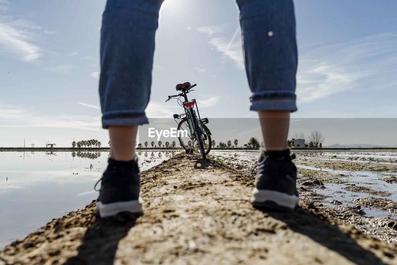 Bicycle seen through legs of woman standing on footpath at ebro's delta during sunny day, spain