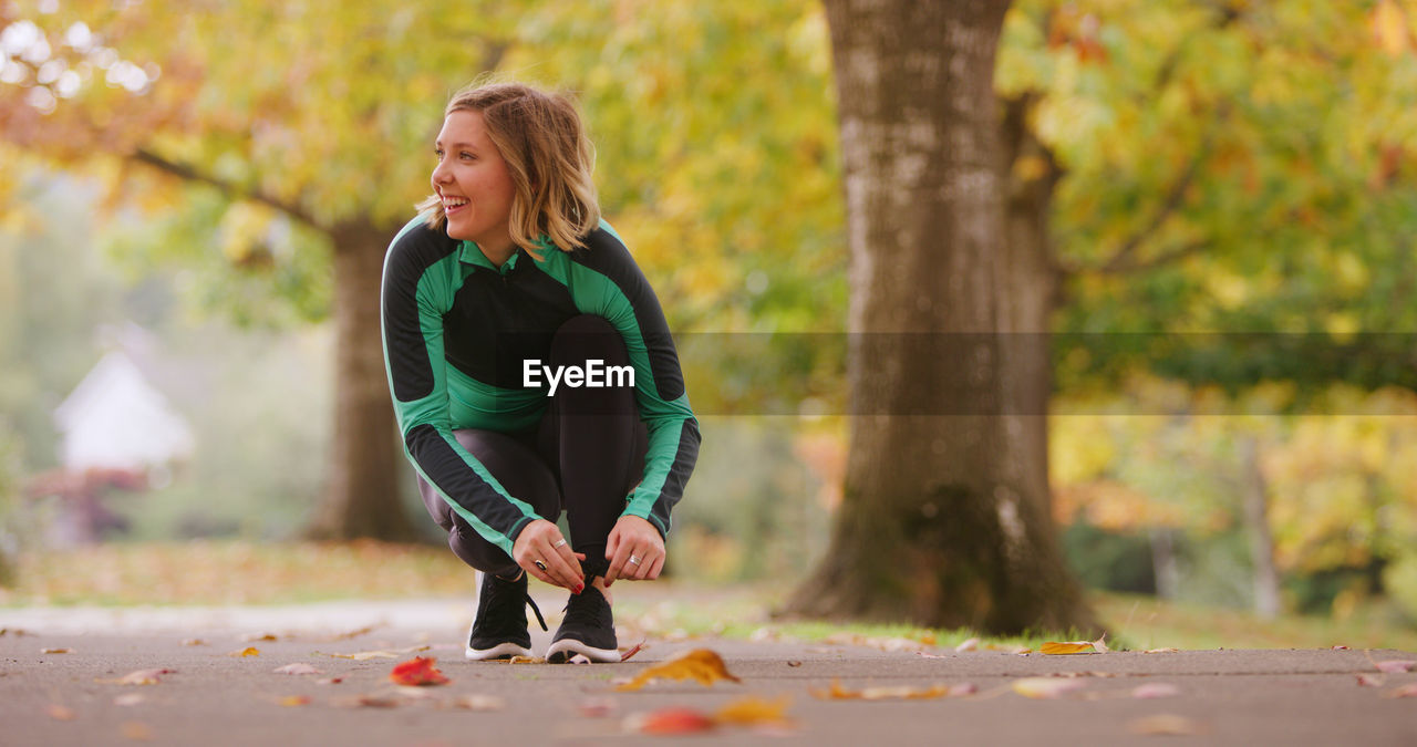 Smiling young woman tying shoelace in park during autumn