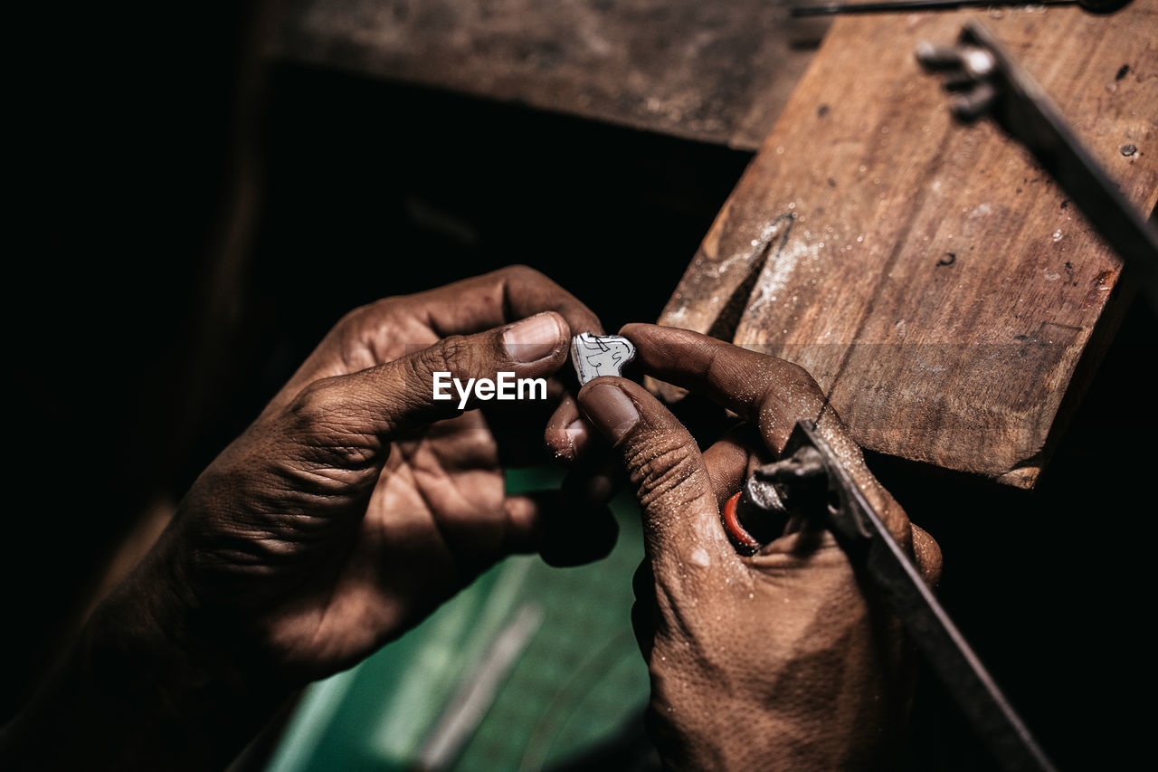 From above faceless craftsman holding metal piercing saw and piece of silver while working at shabby desk