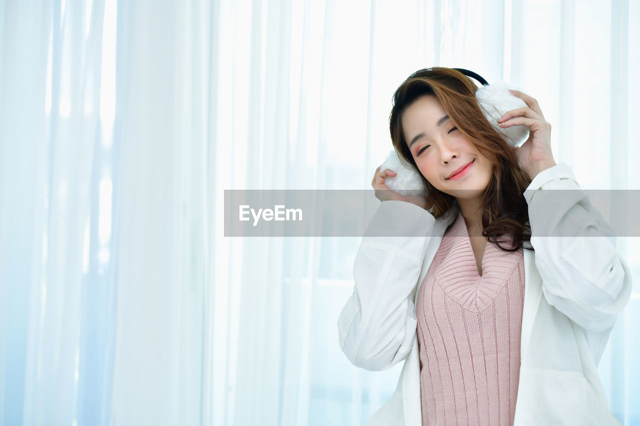 Young woman wearing ear muffs against curtain at home