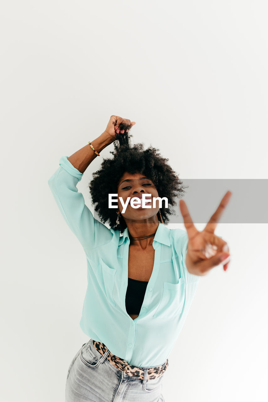 Playful young african american female in trendy outfit having fun and showing tongue and peace sign on white background