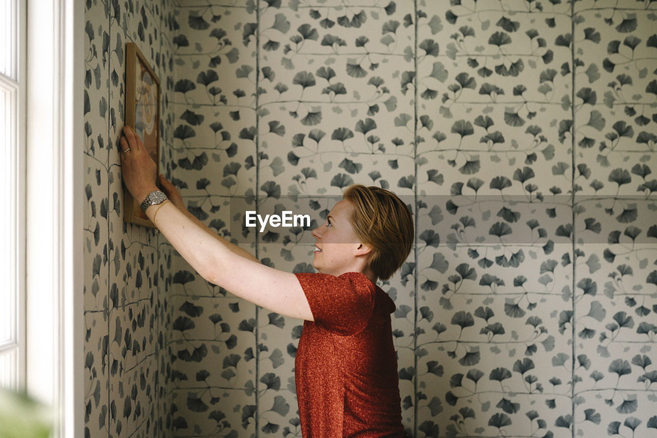 Side view of woman adjusting picture frame on wall at home