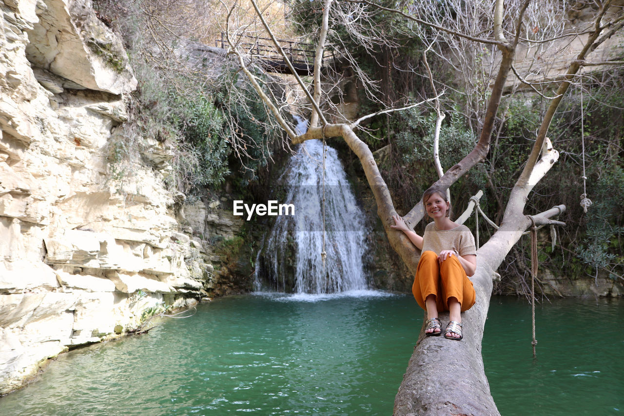 Smiling woman sitting on tree trunk against waterfall