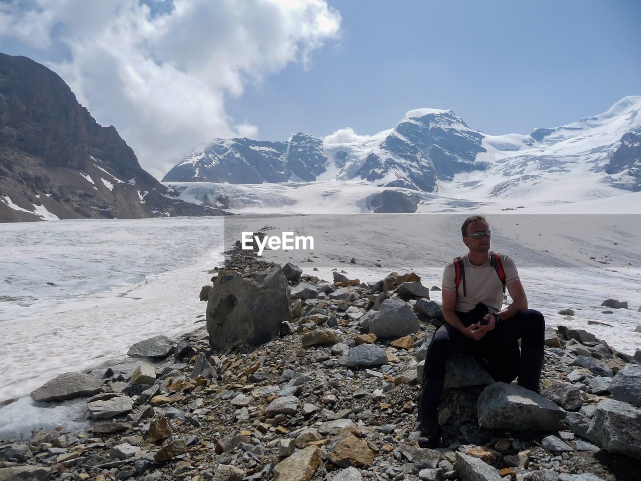 Full length of hiker sitting on rock at snow covered mountain against sky