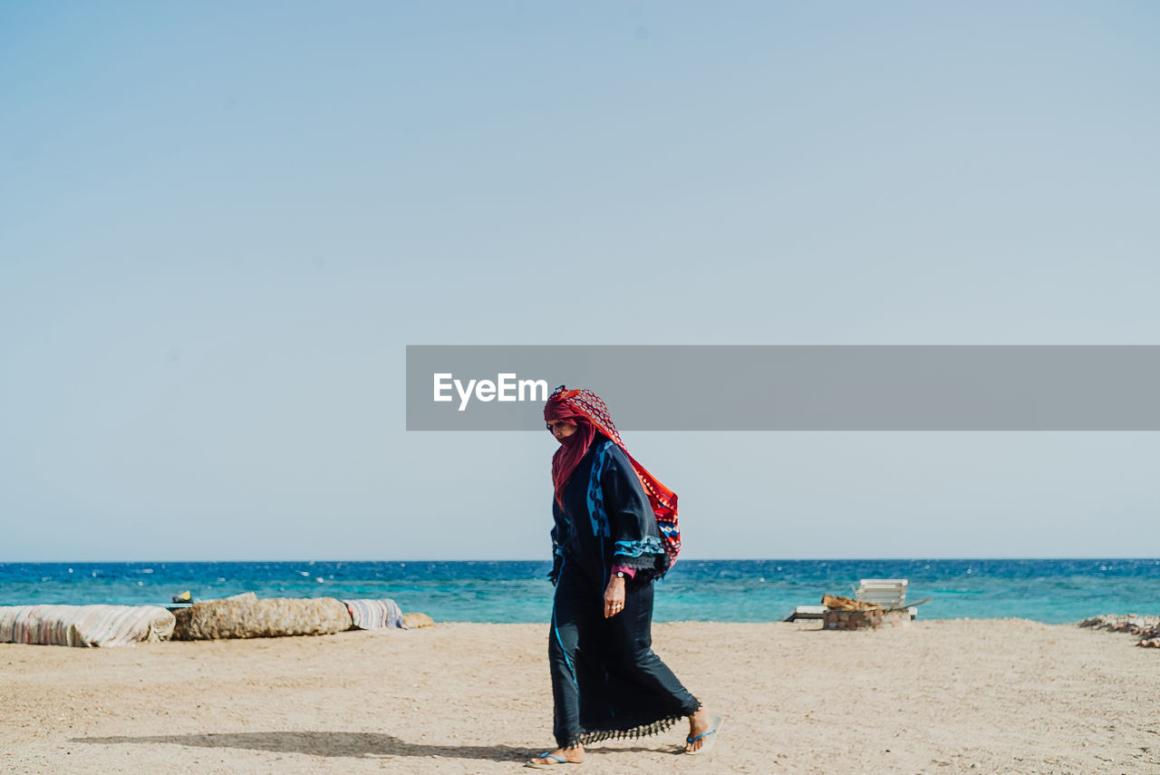 Side view of woman wearing traditional clothing while walking at beach against clear sky