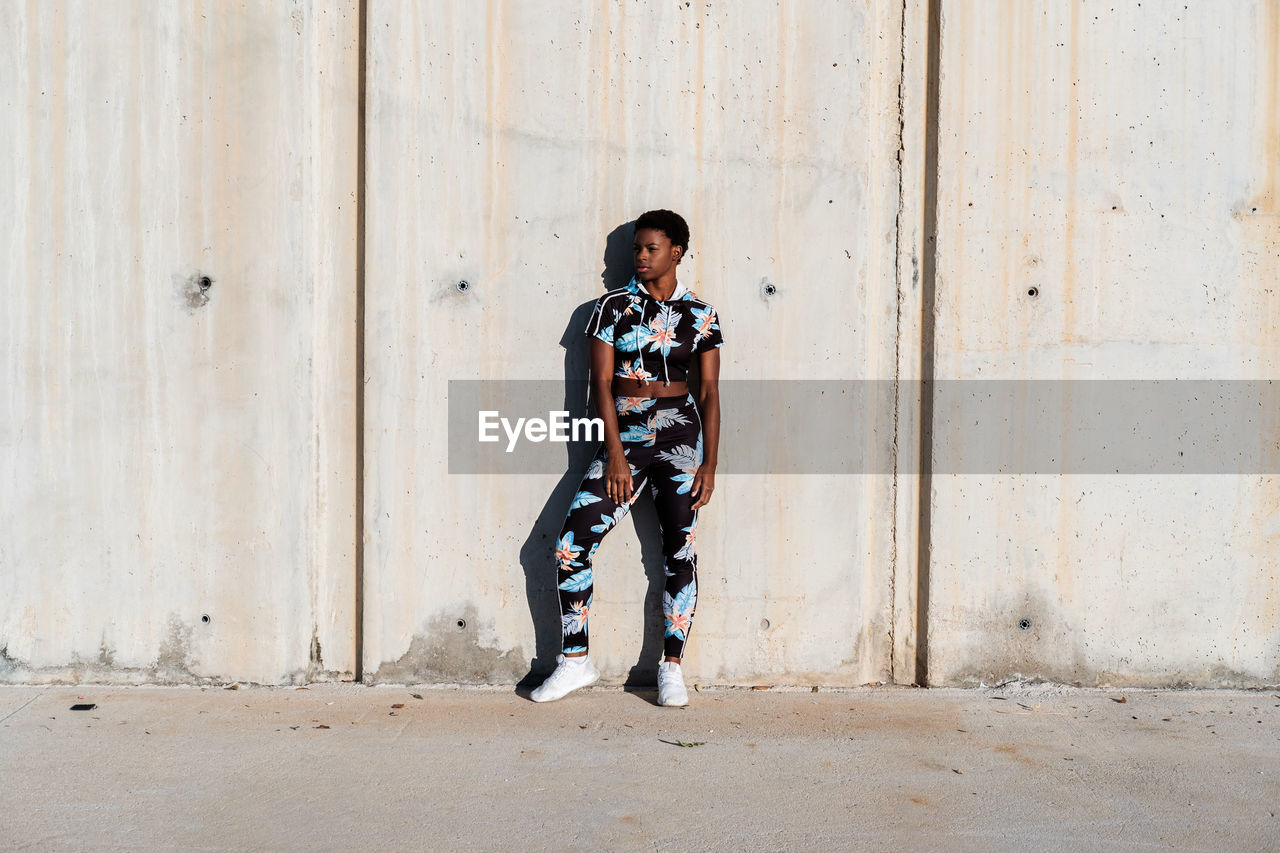 Cheerful confident african american female athlete in flowered sport clothes and white sneakers looking away with interest and laughing while standing alone leaning on concrete wall in sunbeams and resting after training in city