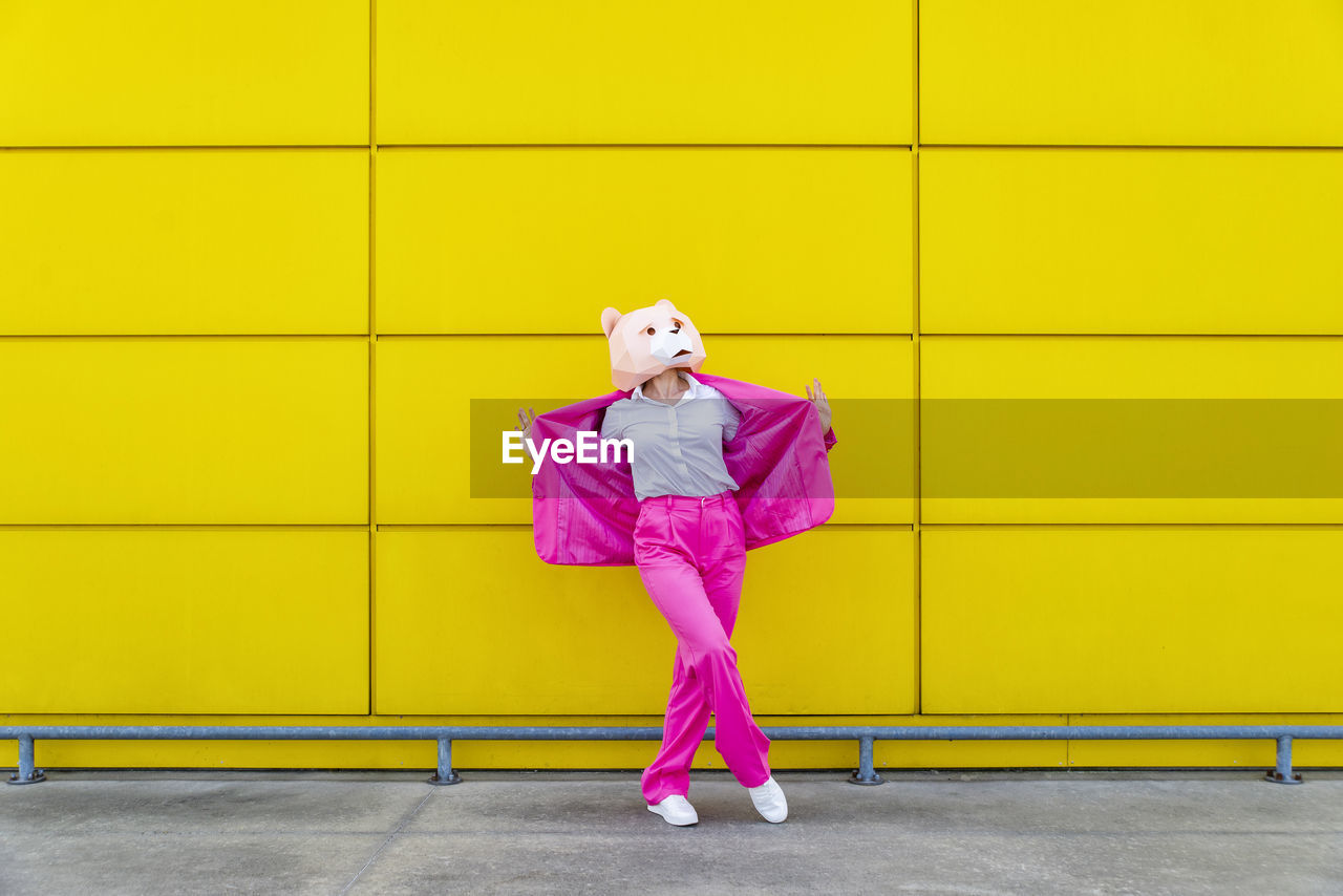 Woman wearing vibrant pink suit and bear mask standing in front of yellow wall with spread jacket