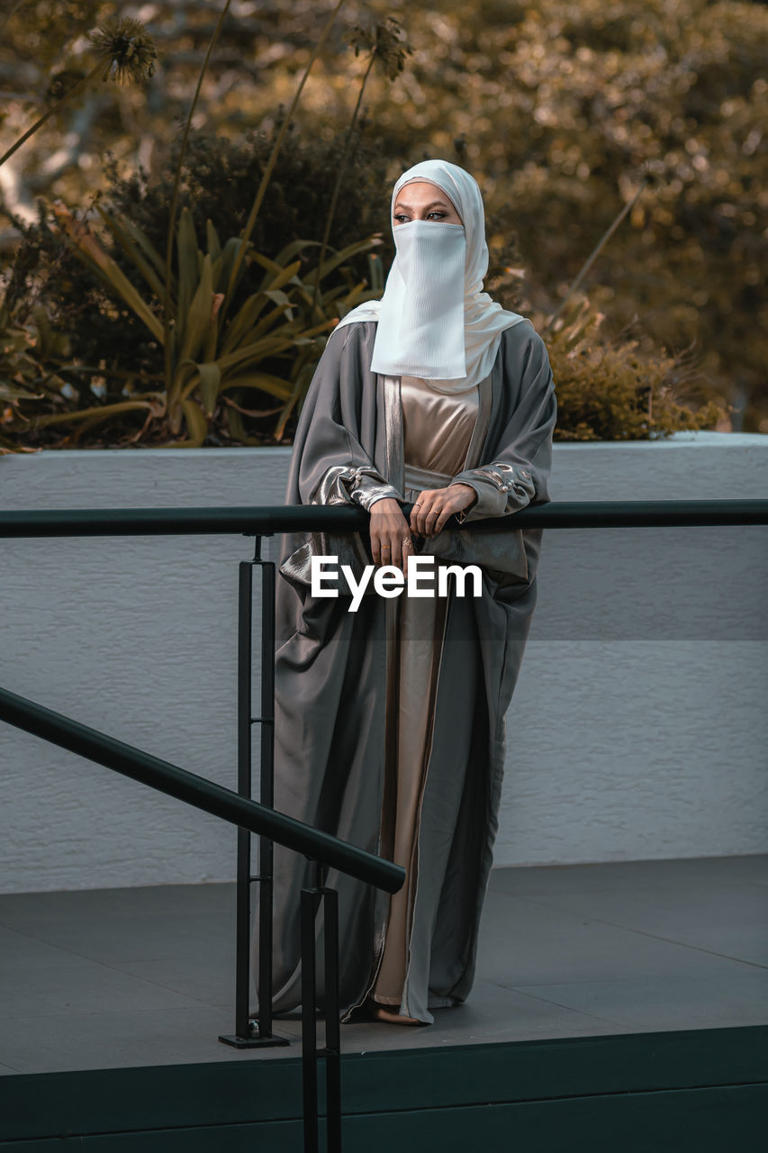 Full length picture of veiled woman with white niqab and hijab standing by railing