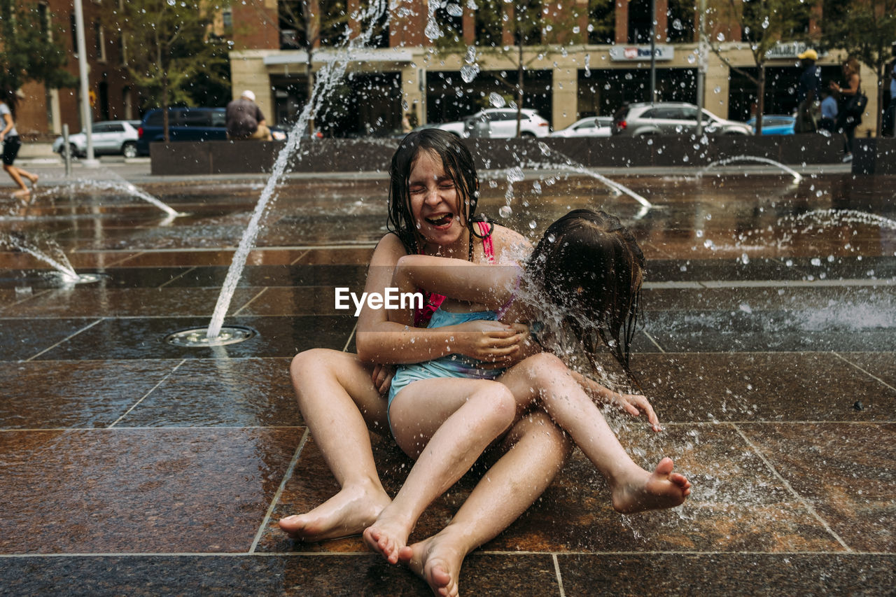 Cheerful sisters wearing swimwear playing with fountain water against buildings in city