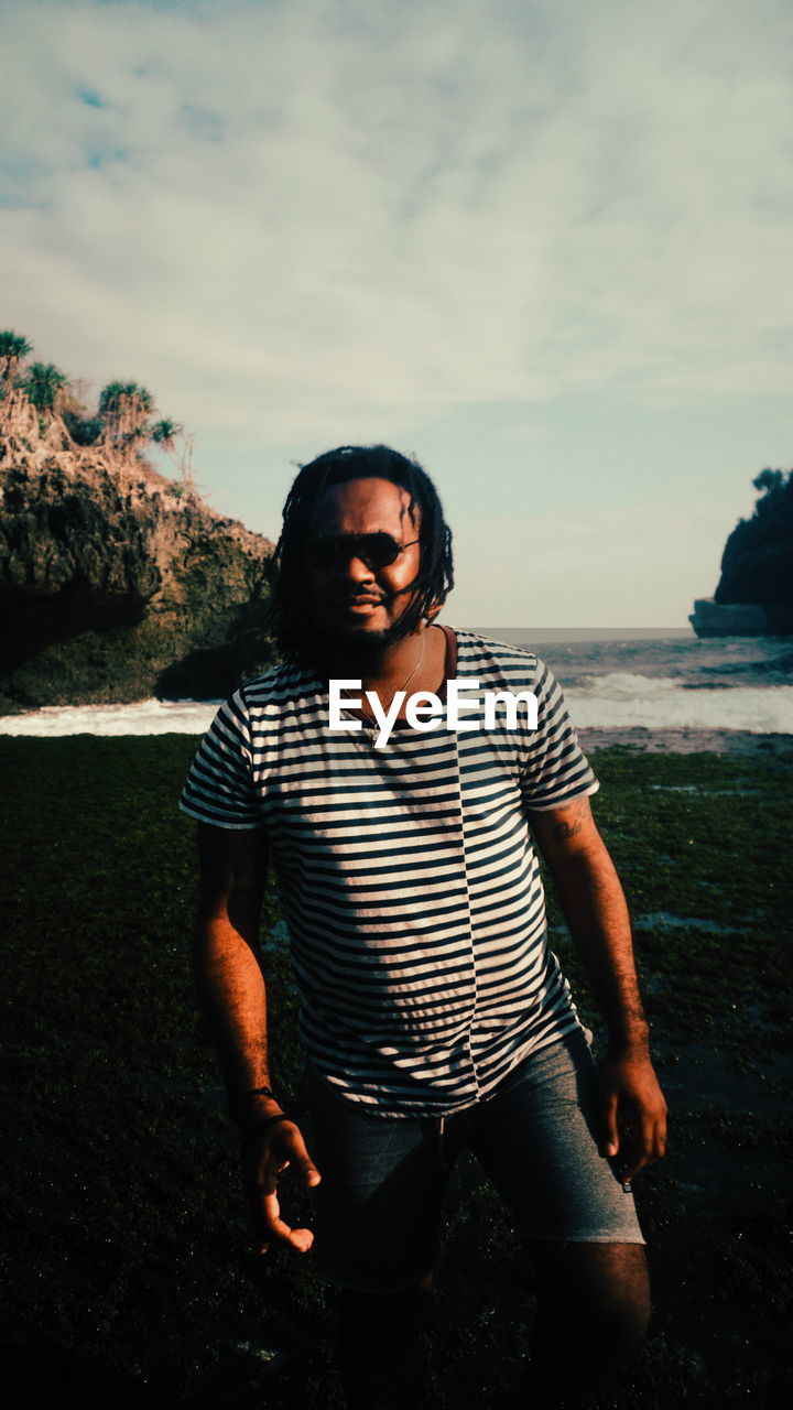 one person, water, sea, sky, nature, beach, land, striped, casual clothing, front view, young adult, men, blue, adult, leisure activity, cloud, portrait, standing, rock, three quarter length, photo shoot, black, outdoors, glasses, beauty in nature, lifestyles, looking at camera, looking, vacation, ocean, sunglasses, cool, day, trip, holiday, relaxation, fashion, person, scenics - nature