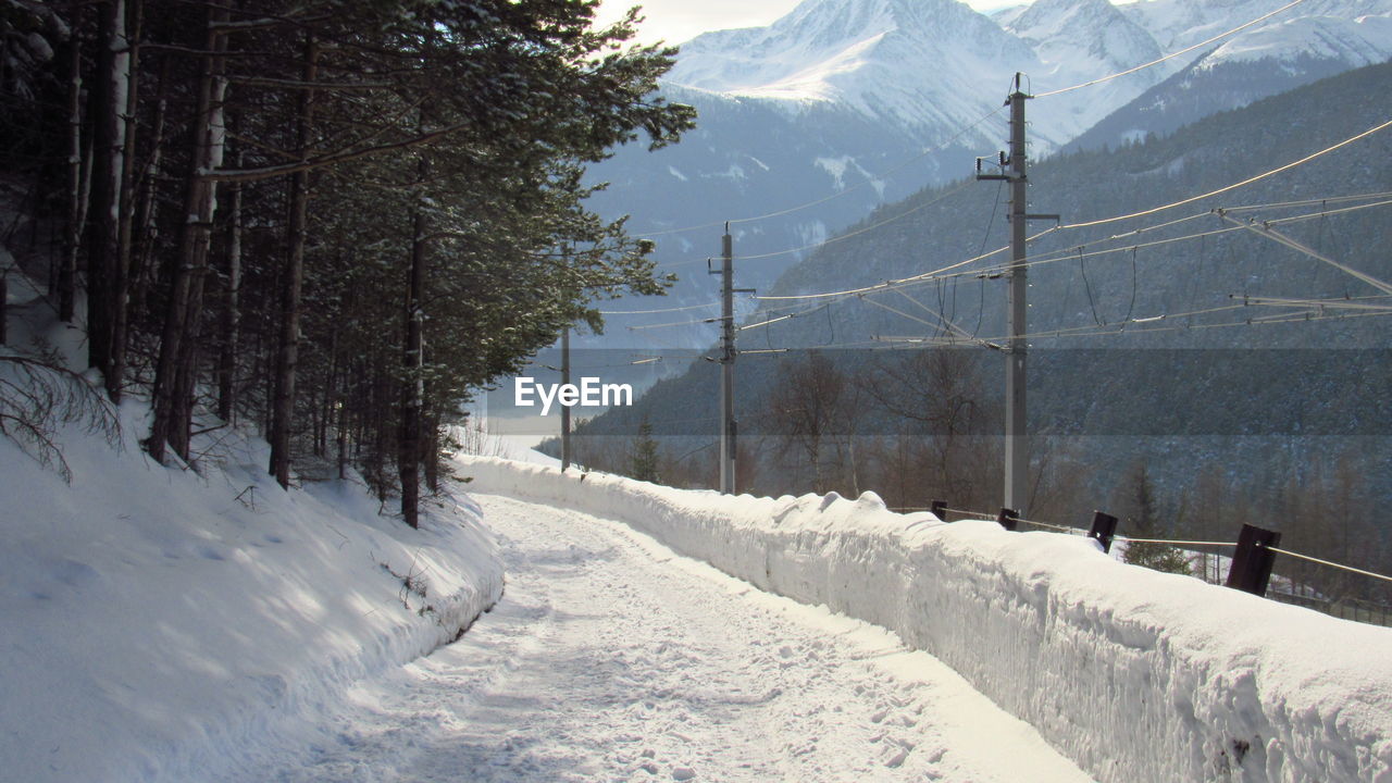 SNOW COVERED ROAD BY TREES AGAINST MOUNTAINS