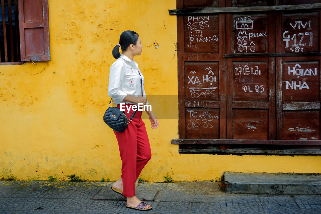 Side view of young woman standing on road against wall