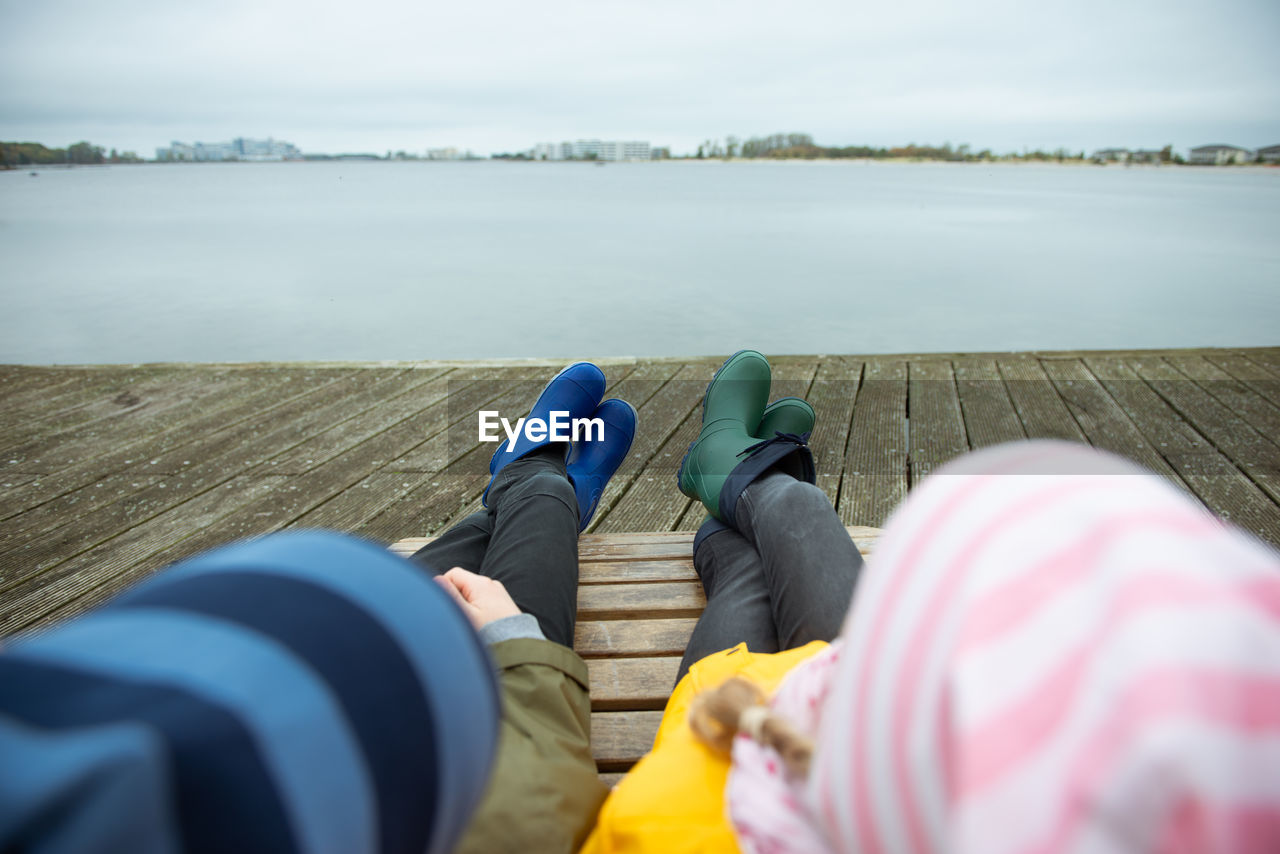 Low section of people relaxing by lake against sky