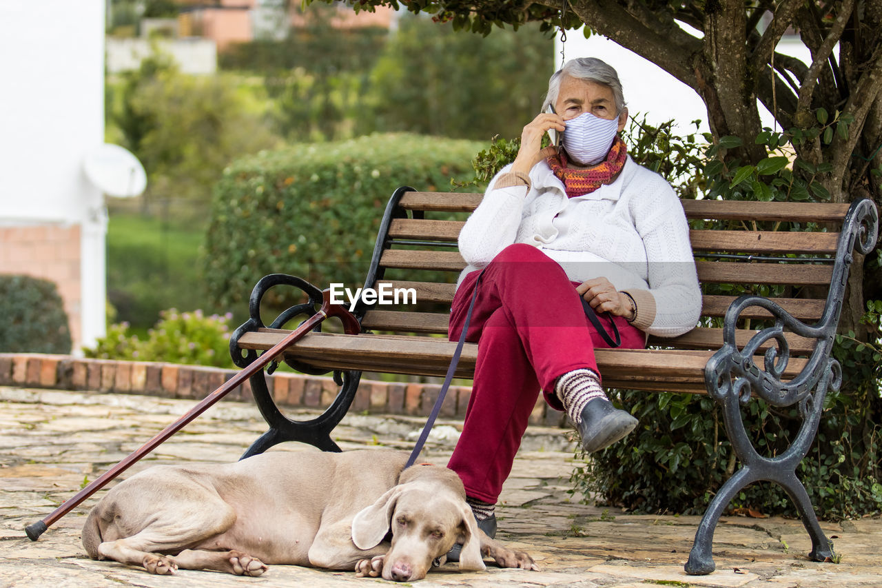 Full length of senior wearing mask with dog sitting outdoors
