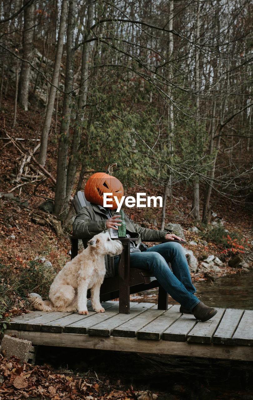 Man wearing scary pumpkin head for halloween sitting on dock with dog.