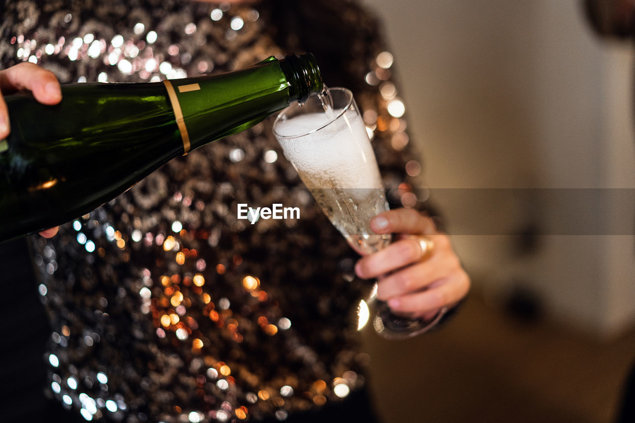 Unrecognizable female in elegant shiny dress standing in room and pouring cold champagne in glass during christmas party