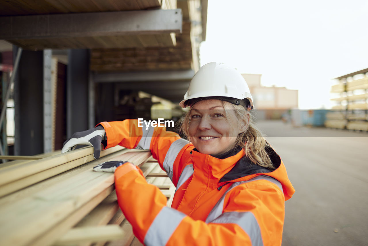 Portrait of smiling female worker in protective workwear stacking planks at industry