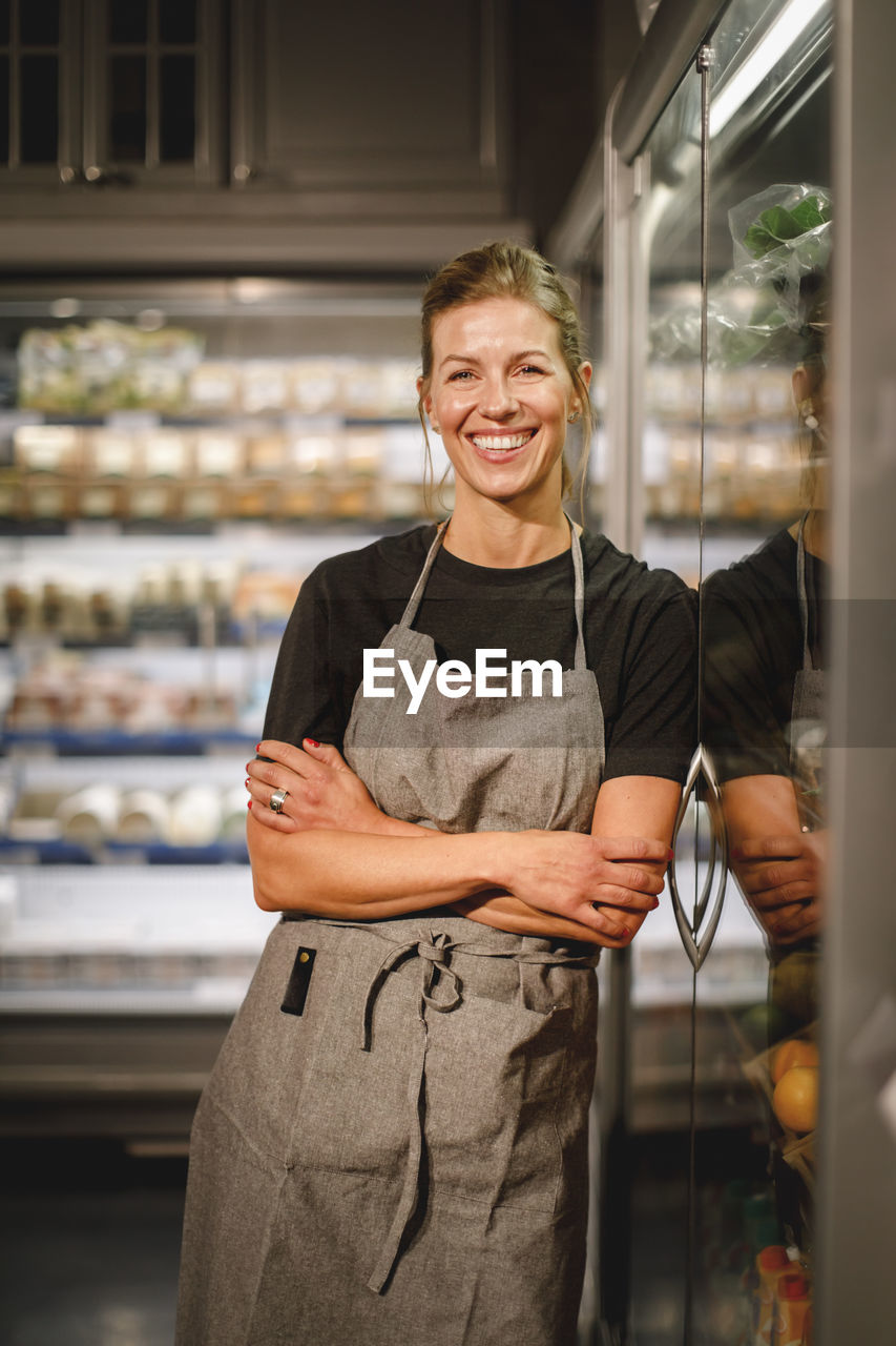 Portrait of smiling saleswoman with arms crossed leaning over refrigerator in grocery store